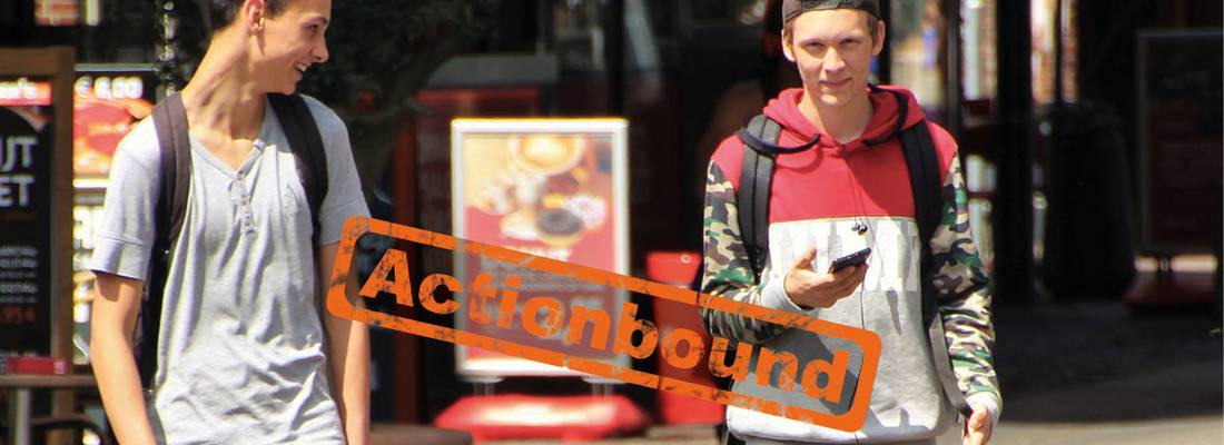 Zwei Jungen mit Handy sind in einer Stadt unterwegs, Stempel Actionbound auf dem Bild
