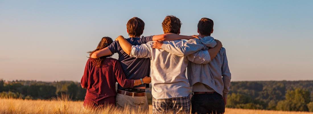 Vier Jugendlichen von hinten fotografiert, sitzen arm in arm auf einem Feld