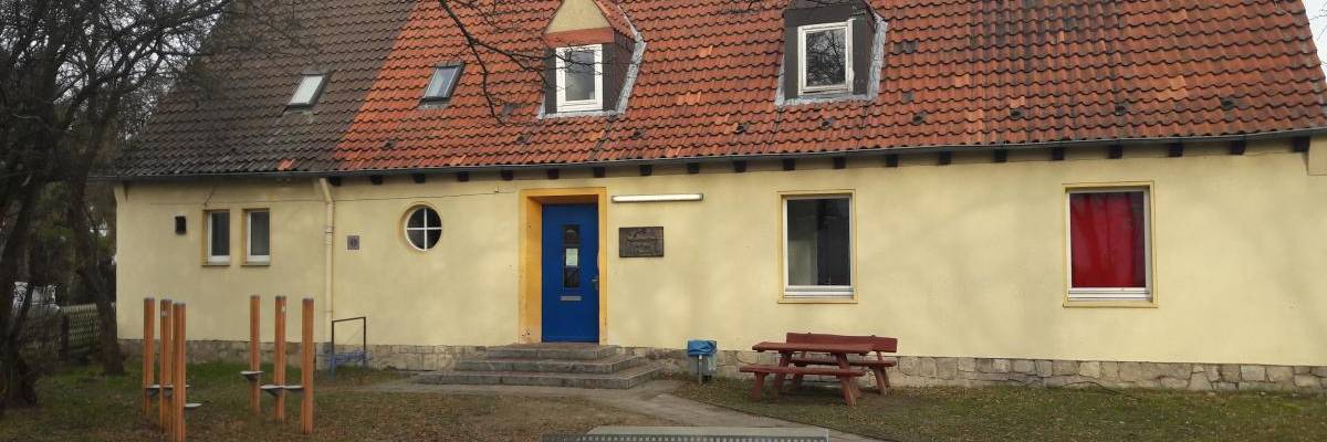 Blick auf die Vorderseite. Gelbes Haus mit rotem Dach und blauer Eingangstür