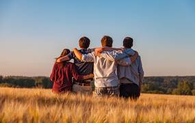 Vier Jugendlichen von hinten fotografiert, sitzen arm in arm auf einem Feld