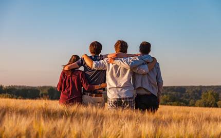 Vier Jugendlichen von hinten fotografiert, sitzen arm in arm auf einem Feld