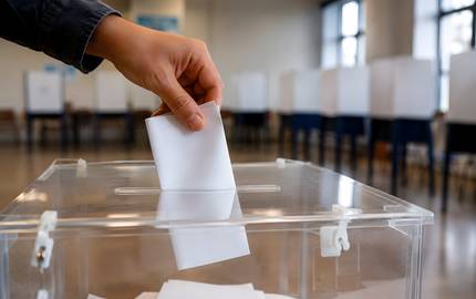 Hand legt Stimmzettel in durchsichtige Wahlurne