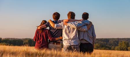 Vier Jugendlichen von hinten fotografiert, sitzen arm in arm auf einem Feld